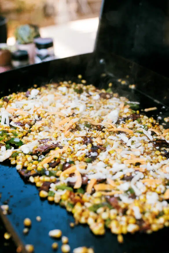 Hatch Chile and Bacon Esquites cooked on a blackstone flattop grill, loaded with cheese and absolutely delicious. Perfect as a game day snack, a topping for tacos, or just an amazing side dish to serve for dinner! 